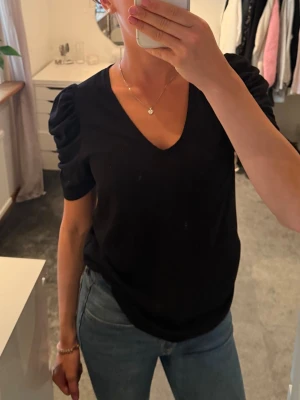Svart topp med puffärm och v-ringning - Snygg svart topp med v-ringad hals och puffade korta ärmar. Toppen har en enkel men trendig design som passar till mycket. Materialet ser ut att vara mjukt och bekvämt, troligen bomull eller polyester. Perfekt för dig som gillar stilrena plagg med en twist.