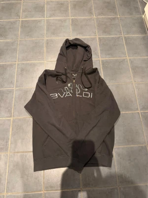 Svart hoodie från Bvaldi med dragkedja - Svart hoodie från Bvaldi med dragkedja framtill och snyggt mönstrad logga i silver på bröstet. Utrustad med huva och svarta dragsnören. Perfekt för en chill och avslappnad stil. Den är använd så den är inte i samma skick som när den köptes. Men där är inga större fel på den! I 