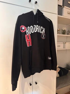 Svart hoodie från Hoodrich med tryck - Svart hoodie från Hoodrich med stora vita och röda tryck och broderade detaljer framtill. Klassisk känguruficka, dragsko i huvan och ribbade muddar. Perfekt för en streetwear-look och riktigt skön att bära.