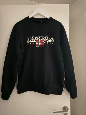 Svart sweatshirt från Napapijri - Cool svart sweatshirt från Napapijri med tryckt logga och norsk flagga på bröstet. Tröjan har rund hals, långa ärmar och ribbade muddar. Perfekt för en avslappnad streetstyle-look.