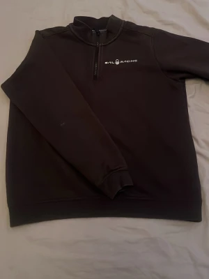 Svart Sail Racing halfzip hoodie - Svart hoodie från Sail Racing med halv dragkedja och logga på bröstet. Tröjan har hög krage, långa ärmar och ribbade muddar. Perfekt för en sportig och stilren look. Materialet känns mjukt och bekvämt mot huden.