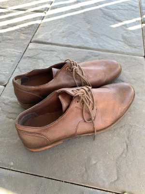 Bruna klassiska loafers  - Snygga bruna loafers med snörning och låg klack. Skorna har en stilren design med rundad tå och diskreta sömmar. Perfekta för dig som gillar en tidlös och clean look. Passar till både jeans och chinos.