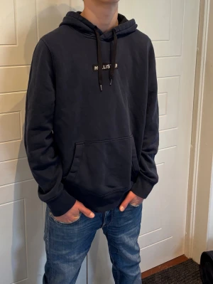 Mörkblå hoodie från Hollister - Snygg mörkblå hoodie från Hollister med klassisk känguruficka och justerbar huva med svarta snören. Diskret Hollister-logga på bröstet. Tillverkad i mjukt bomullsmaterial som känns skönt mot huden. Perfekt för en avslappnad och stilren look. Passar S