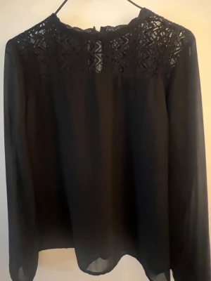 Svart blus med spetsdetaljer - Snygg svart blus med transparenta långärmar och vacker spets upptill. Blusen har en lös passform och rund halsringning med fina mönster i spetsen. Perfekt för dig som gillar stilrena och trendiga plagg.