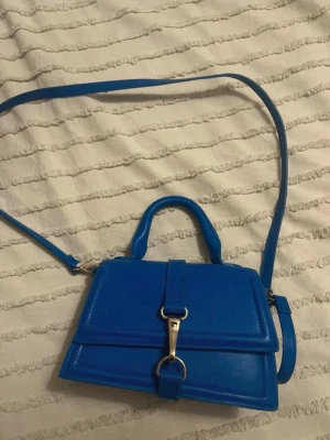 Blå axelremsväska med silverdetalj - Snygg blå handväska i slätt konstläder med både handtag och justerbar axelrem. Väskan har en stilren design med silverfärgad spänndetalj framtill och lockstängning. Perfekt för dig som vill ha en färgklick till din outfit.