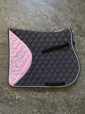 Brun och rosa schabrak från EQuest - Säljer ett snyggt schabrak från EQuest i brunt med ljusrosa detalj och broderad logga. Schabraket har rutmönster, vita kanter och svarta remmar för fäste. Perfekt till ridning och passar de flesta sadlar.