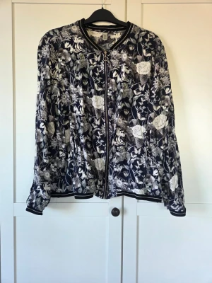 Blommig bomberjacka i mörka toner - Snygg bomberjacka med svart botten och blommigt mönster i vitt och grått. Jackan har dragkedja framtill, ribbade muddar vid ärmslut och nederkant samt klassisk bomberkrage. Perfekt för dig som vill sticka ut med en unik och trendig look.