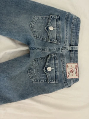 True Religion Joey ljusblå jeans - Säljer ett par ljusblå jeans från True Religion med ikoniska ficklock och vita knappar bak. Modell: Joey. Jag har sytt upp dem en liten bit så att det passar min längd, 165 cm.