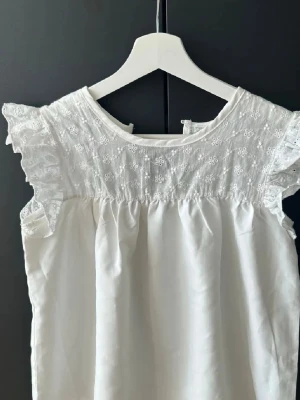 Vit blus med spets och volangärmar - Supersöt vit blus med broderad spets upptill och volangärmar. Blusen har rund halsringning och små blommiga detaljer i spetsen. Perfekt för en fräsch och romantisk look.