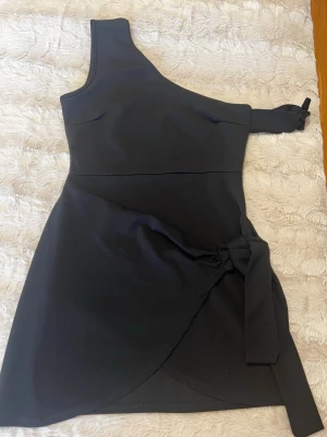 Svart oneshoulder klänning med knyt - Snygg svart kortklänning med oneshoulder-design och knyt i midjan. Klänningen har omlott-känsla nertill och en ärm med rosett-detalj. Perfekt för dig som vill sticka ut med en modern och stilren look.