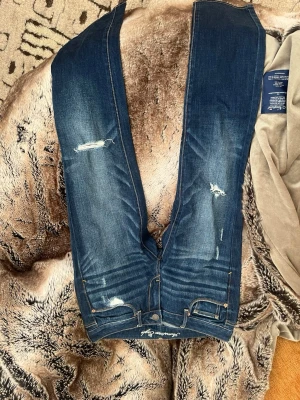 Mörkblå slitna jeans American Eagle - Säljer ett par mörkblå jeans från American Eagle med snygga slitningar och skinny fit. Jeansen har klassisk femficksdesign, gylf med knapp och dragkedja samt kontrastsömmar. Perfekta för en trendig och avslappnad look.