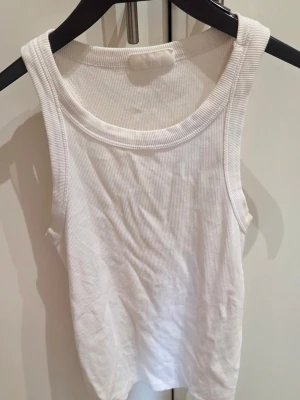Vit ribbad linnetopp från Gina Tricot - Säljer en klassisk vit ribbad linnetopp från Gina Tricot, basically basics-kollektionen. Toppen är ärmlös med rund halsringning och har en enkel, stilren design som passar till allt. Perfekt att styla lager på lager eller som den är.