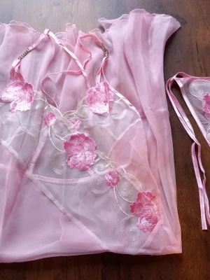 Rosa spetsbody med blommor storlek S - Säljer ett set med en rosa body och matchande trosa i transparent mesh och spets. Bodyn har tunna axelband och är dekorerad med broderade blommor i rosa och vitt. Materialet är lätt och luftigt, perfekt för dig som gillar romantiska detaljer. Storlek S 