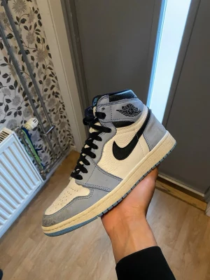 Nike Air Jordan 1 High blå/vit - Snygga Nike Air Jordan 1 High sneakers i vitt och ljusblått läder med svarta detaljer och klassisk Swoosh. Högt skaft, svart logga på sidan och blå yttersula. Perfekta för dig som vill sticka ut med stilrena och ikoniska sneakers.