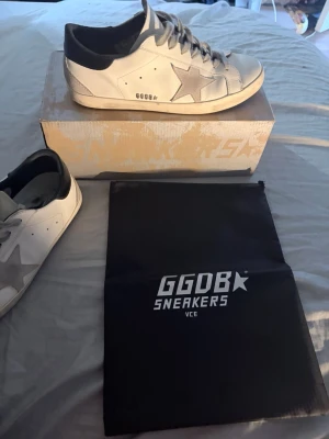 Golden Goose vita sneakers med stjärna - Golden Goose sneakers i vitt läder med grå mockastjärna på sidan, grå skosnören och svart hälparti. Klassisk låg modell med GGDB-logga och slitna detaljer för en cool streetlook. Kommer med originalpåse och box.