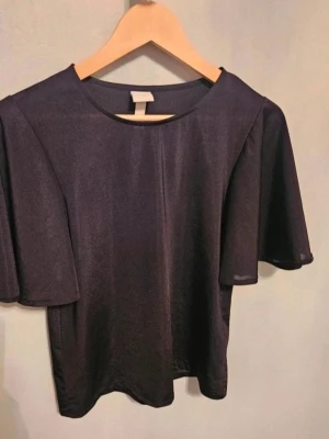 Topp H&M XS - Svart glansig topp med vida ärmar H&M XS  Säljer en svart glansig topp från H&M i storlek XS. Toppen har vida, korta ärmar och en rak passform. Materialet är tunt och skimrande, vilket ger en snygg look. Perfekt för dig som gillar stilrena plagg med det lilla extra.  Kan även hämtas I Partille  E1