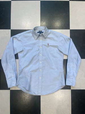 Ljusblå skjorta från Ralph Lauren - Klassisk ljusblå skjorta från Ralph Lauren med button-down krage och broderad logga på bröstet. Skjortan har långa ärmar, knappar framtill och är tillverkad i mjuk bomull. Perfekt för en clean och stilren look.