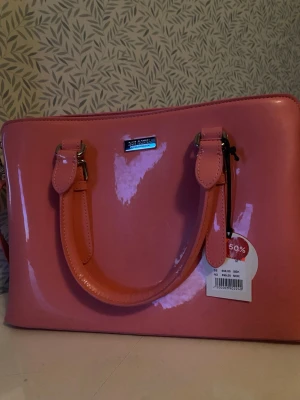 Rosa lackad handväska Don Donna - Säljer en glansig rosa handväska från Don Donna med två stadiga handtag och silverfärgade metalldetaljer. Väskan har en rektangulär form och är tillverkad i lackat material som ger en snygg och modern look. Perfekt för dig som vill sticka ut med färg.