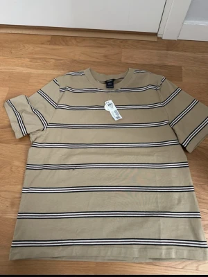 Beige randig t-shirt från Lindex - Snygg beige t-shirt från Lindex med svarta och vita horisontella ränder. Klassisk passform med korta ärmar och rund halsringning. Perfekt för en avslappnad och trendig stil.