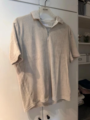 Beige frottétopp med krage och zip - Beige topp i mjuk frotté med kort ärm, krage och halv dragkedja framtill. Perfekt för en chill och avslappnad stil. Enkel och stilren design utan mönster eller logga.