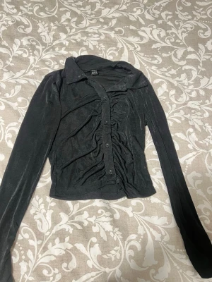 Svart sammetskjorta från SHEIN - Snygg svart skjorta från SHEIN i sammetsmaterial med långa ärmar och knappar framtill. Skjortan har en cool rynkad detalj på framsidan som ger en unik look. Perfekt för dig som vill sticka ut lite extra.
