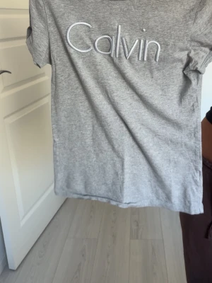 Grå Calvin Klein t-shirt - Snygg grå t-shirt från Calvin Klein med stort broderat 'Calvin' på bröstet. Klassisk passform och korta ärmar, perfekt till jeans eller shorts. 
