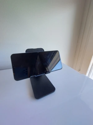Vikbart mobiltelefonställ för skrivbord - Praktiskt svart mobilställ i plast som passar de flesta smartphones. Stället har en enkel och stilren design och kan justeras för både liggande och stående läge. Perfekt för att kolla på videos eller ha mobilen synlig på skrivbordet.