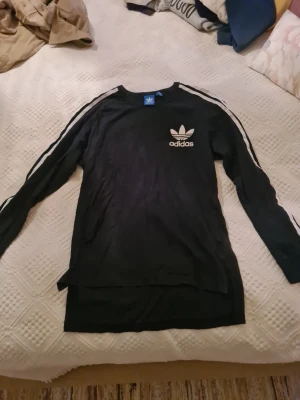 Svart långärmad Adidas tröja - Svart långärmad tröja från Adidas med vita ränder längs ärmarna och klassisk Adidas-logga på bröstet. Tröjan har rund halsringning och är tillverkad i mjukt material. 