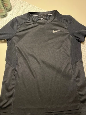 Svart Nike löpartröja - Snygg svart löpartröja från Nike med korta ärmar och rund hals. Tillverkad i ett lätt och ventilerande funktionsmaterial som passar perfekt till träning. Diskret Nike-logga på bröstet och meshpaneler för extra andningsförmåga.