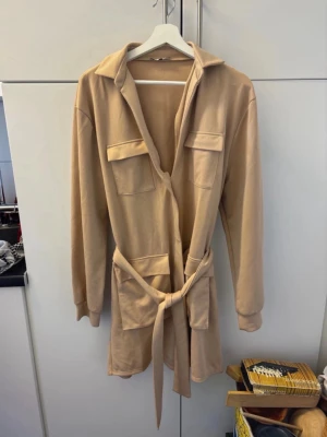 Beige klänning med bälte - Stilren beige tklänning med långa ärmar, krage och två stora bröstfickor. Klänningen har ett knytbälte i midjan för en snygg siluett och är tillverkad i ett mjukt material som känns skönt mot huden.