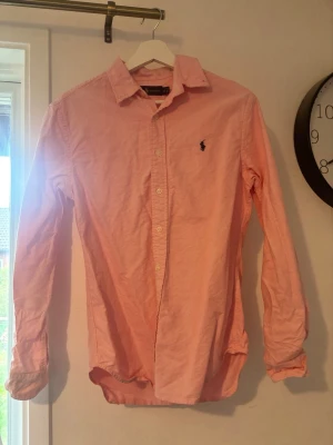 Rosa skjorta från Ralph Lauren - Säljer en klassisk rosa skjorta från Ralph Lauren med button-down krage och broderad logga på bröstet. Skjortan har lång ärm och knäppning framtill. Perfekt för dig som gillar stilrena och tidlösa plagg.