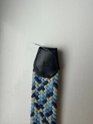 Flätat bälte från ONEWear i blå toner - Snyggt flätat bälte från ONEWear i olika blå nyanser och beige. Bältet har detaljer i svart skinn och ett klassiskt spänne i metall. Perfekt accessoar för att lyfta din outfit med lite färg och stil.