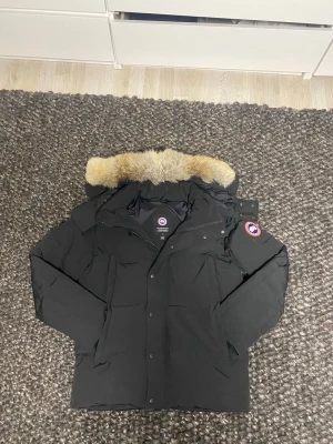 Svart dunjacka från Canada Goose - Säljer en svart dunjacka från Canada Goose med avtagbar huva och äkta päls på kragen. Jackan har klassisk logga på ärmen, dragkedja och knappar framtill samt flera fickor. Perfekt för kalla vinterdagar.