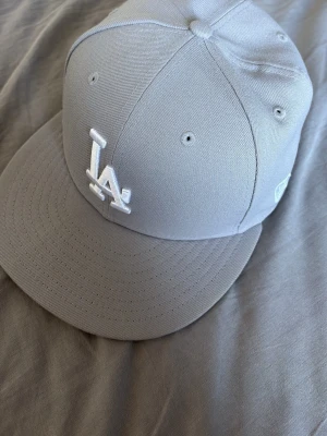 Grå LA-keps från New Era 59FIFTY - Snygg grå keps från New Era 59FIFTY med LA-logga broderad framtill i vitt. Klassisk flat brim och strukturerad passform. Kepsen är tillverkad i ett slitstarkt tyg och har broderade ventilationshål för extra komfort.