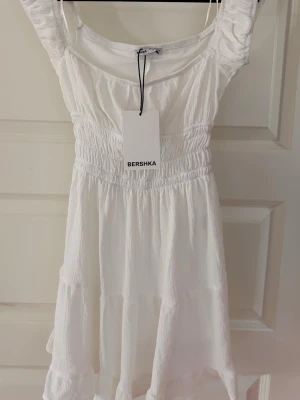 Klänning från Bershka - Söt vit kort klänning från Bershka med smockad midja och volangdetaljer. Klänningen är aldrig använd. Har även inbydda shorts.💕
