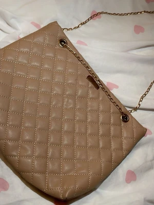 Beige quiltad handväska med kedja - Stilren beige handväska i quiltat mönster med guldfärgad kedjerem. Väskan har en mjuk, rektangulär form och är tillverkad i konstläder. Perfekt accessoar för att lyfta din outfit med en trendig touch.