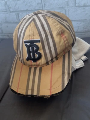 Beige keps med Burberry-mönster - Snygg keps från Burberry i klassiskt beige med bruna, svarta och röda ränder. Framtill finns ett stort svart TB-logotypbroderi. Kepsen har böjd skärm och justerbar rem baktill. Perfekt för en trendig streetstyle-look.