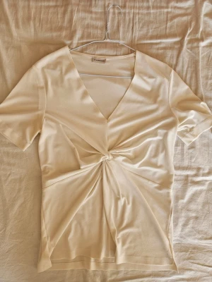 Topp från By Malene Birger - Snygg vit topp från By Malene Birger med v-ringning och en fin detalj framtill. T-shirtrmad och i ett mjukt, lätt material som känns lyxigt. Perfekt för dig som gillar stilrena och trendiga plagg.