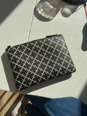 Svartvit kuvertväska med mönster - Snygg svartvit kuvertväska med grafiskt rutmönster och dragkedja upptill. Väskan är i ett tåligt material som känns som konstläder och har guldfärgade metalldetaljer. Perfekt att bära till både jeans och klänning för en trendig look.
