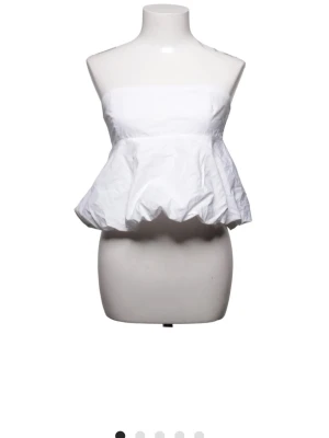 Vit axelbandslös tubtopp med volang - Snygg vit axelbandslös tubtopp med volang och ballongformad nederdel. Toppen har en bandeau-stil och smockad rygg för extra komfort. Perfekt för dig som vill ha en trendig och luftig look.
