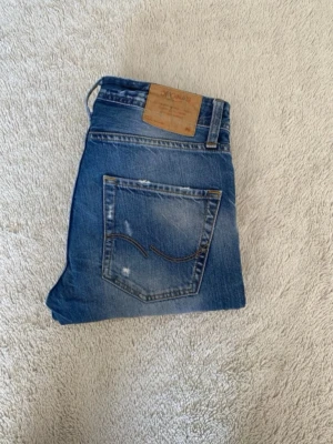 Blå slim fit jeans från Jack & Jones - Säljer ett par blå slim fit jeans från Jack & Jones med slitningar och ljusa detaljer på benen. Jeansen har klassisk femficksdesign, snyggt läderpatch bak och är tillverkade i bomull. Perfekt för dig som gillar en trendig och avslappnad look.