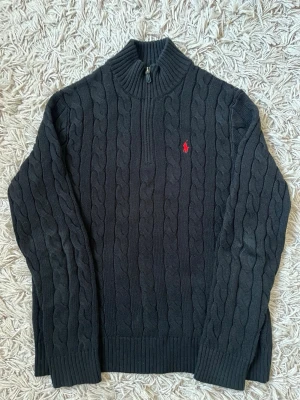 Polo Ralph Lauren svart cableknit - Kabelstickad svart tröja från Polo Ralph Lauren med halv dragkedja och röd broderad logga på bröstet. Tröjan har hög krage, långa ärmar och ribbade muddar. Perfekt för dig som gillar klassisk och stilren design.