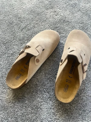 Sand Birkenstock Boston clogs  - Säljer ett par klassiska Birkenstock Boston tofflor i beige mocka med justerbart spänne i metall. Sköna innersulor med soft footbed och en stilren design som passar perfekt till jeans eller mjukisbyxor. Ikonisk modell från Birkenstock, tillverkad i Tyskland. Använda 3 gånger 