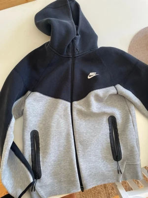 Nike hoodie svart och grå M - Nike hoodie i svart och ljusgrått med dragkedja och två fickor framtill med svarta detaljer. Klassisk Nike-logga på bröstet. Mjuk och bekväm, perfekt för chill eller träning. Huva och långa ärmar. Materialet är en bomullsblandning.