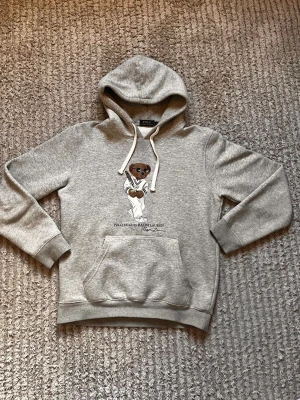 Grå hoodie Polo Bear  - Grå hoodie från Ralph Lauren med Polo Bear-tryck på bröstet. Klassisk modell med huva, vita dragsnören och stor magficka. Mjuk bomullsblandning och ribbade muddar vid ärmslut och nederkant. Perfekt för en avslappnad och trendig stil. Tröjan är i L men passar M. Hör av dig vid fler funderingar!
