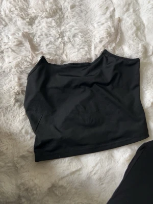 Svart linnetopp med spetsdetalj - Svart linnetopp med tunna axelband i vit spets. Toppen har en enkel och stilren design med rak passform och ärmlös modell. Perfekt att matcha med jeans eller kjol för en trendig look.