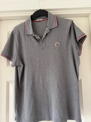 Grå pikétröja från Moncler - Snygg grå pikétröja från Moncler med klassisk krage och knappar framtill. Tröjan har Moncler-logga på bröstet och rödvita detaljer på krage och ärmslut. Perfekt för en stilren och sportig look.