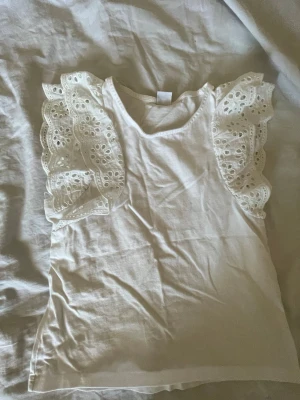 Vit topp med volangärmar - Superfin vit topp med breda volangärmar i broderad spets. Ärmlös modell med rund halsringning och romantiska detaljer. Perfekt för dig som vill ha en söt och trendig look.