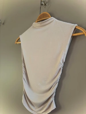 Beige ärmlös topp med rynkade sidor - Snygg beige ärmlös topp med hög hals och rynkade sidor. Toppen har en clean och stilren look, perfekt att matcha med jeans eller kjol. Mjuk och stretchig känsla i materialet för extra komfort.