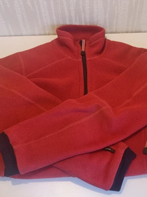 Röd fleecejacka från Haglöfs - Mysig röd fleecejacka från Haglöfs med dragkedja framtill. Jackan har svarta detaljer vid ärmsluten och är tillverkad i mjukt fleecematerial, perfekt för kyliga dagar och outdooraktiviteter. Katt finns i hemmet.